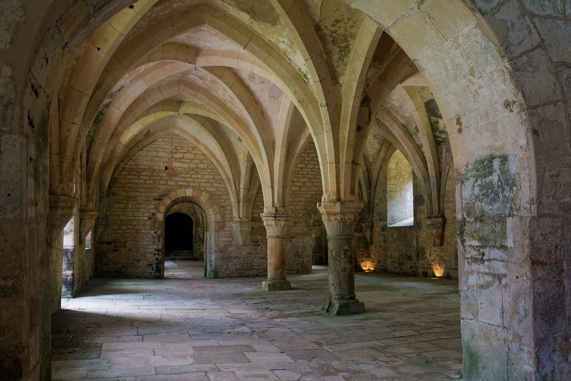 Salle voûtée d’ogives sur fines colonnes à chapiteaux, retombées sur des piliers ronds centraux, sol dallé et murs de pierre nue, à l’abbaye de Fontenay
