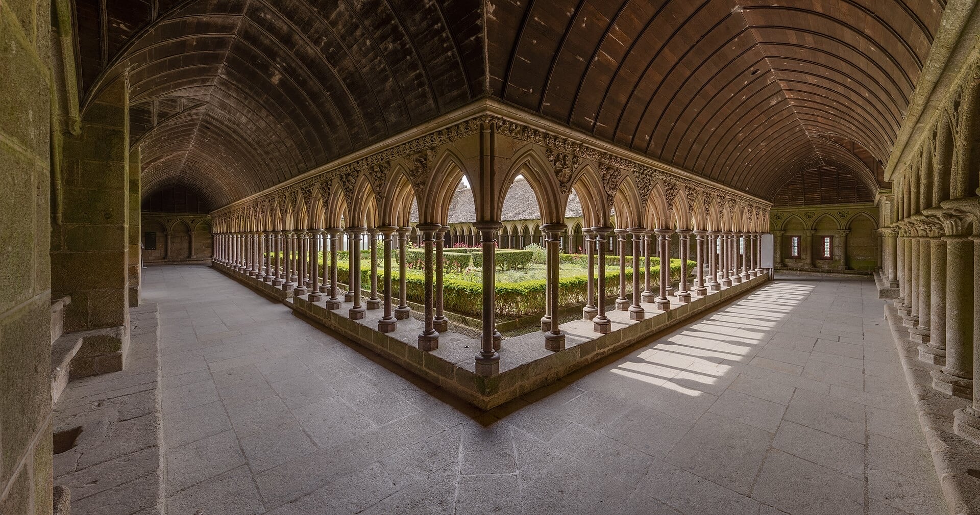 Cloître gothique du Mont-Saint-Michel : doubles rangs de fines colonnettes en quinconce portant des arcades brisées finement sculptées, plafond de bois lambrissé, jardin central