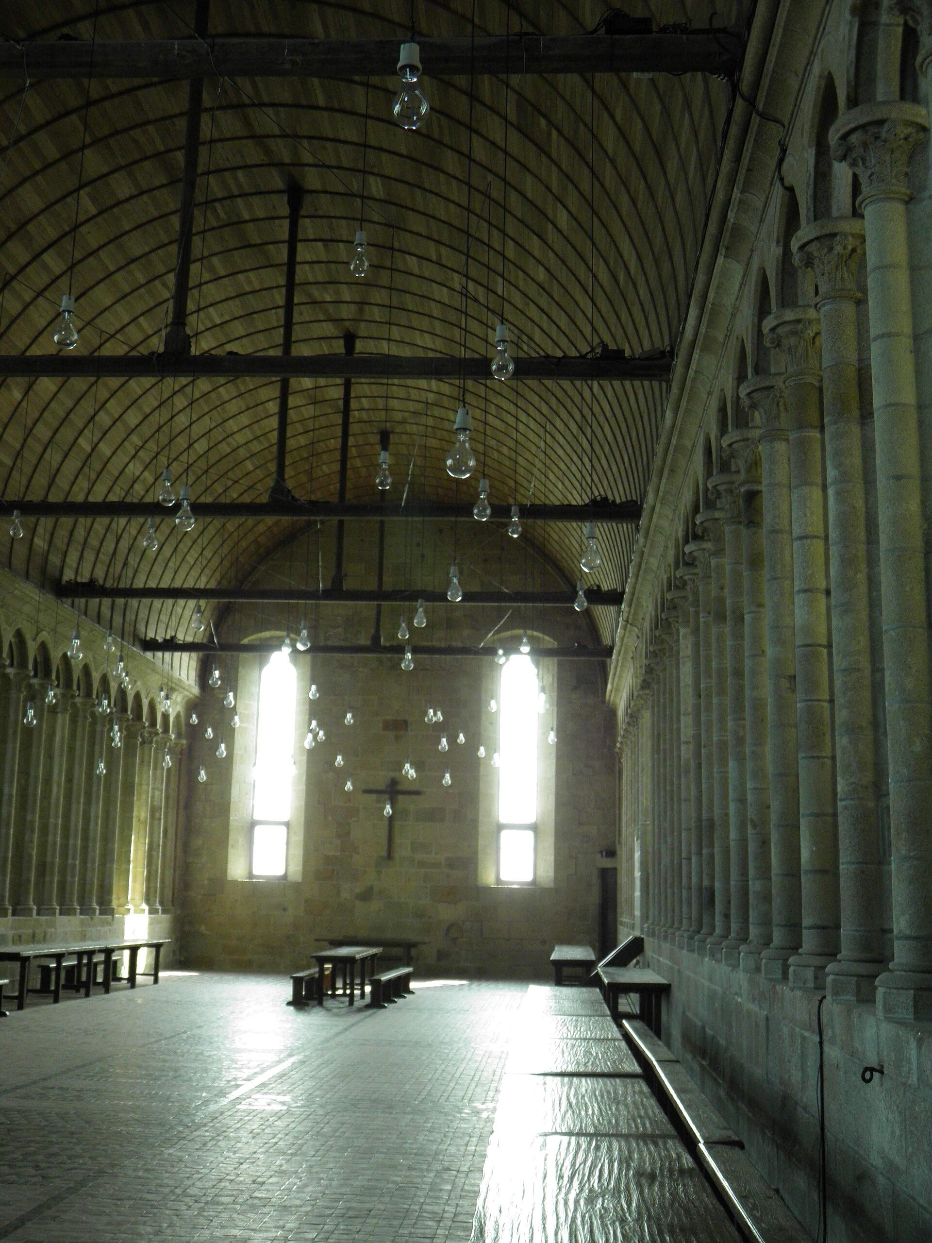 Réfectoire du Mont-Saint-Michel : longue salle à haute voûte de bois en berceau, murs percés d’une multitude de fenêtres étroites et hautes en profonds ébrasements, tables et bancs, crucifix au fond