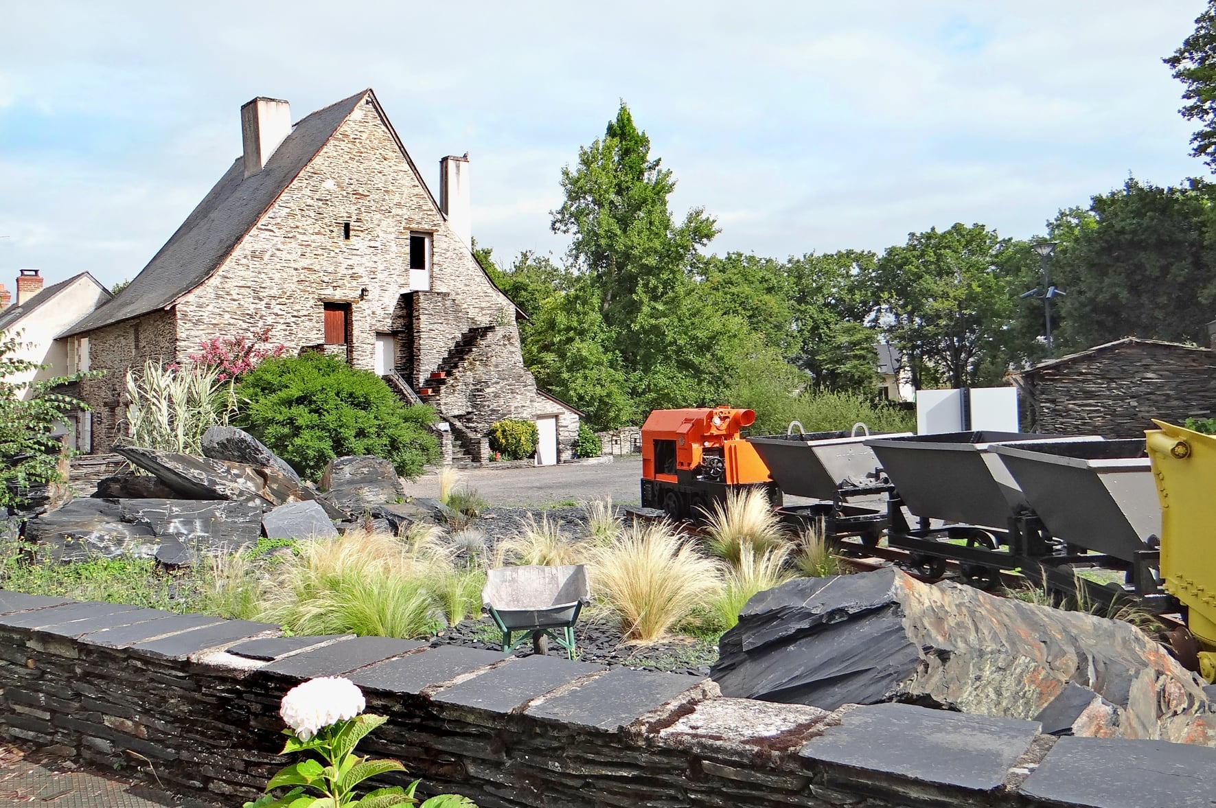 Le musée de l’Ardoise à Trélazé, en Anjou : blocs de schiste ardoisier brut, wagonnets et locotracteur d’extraction exposés devant un ancien bâtiment de moellon de schiste couvert d’ardoise.