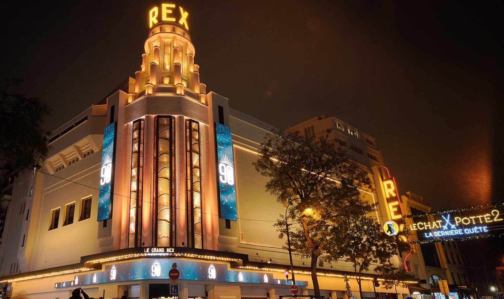 La façade du cinéma Le Grand Rex illuminée la nuit : haute tour d’angle Art déco à gradins portant l’enseigne dorée « Rex », marquise lumineuse