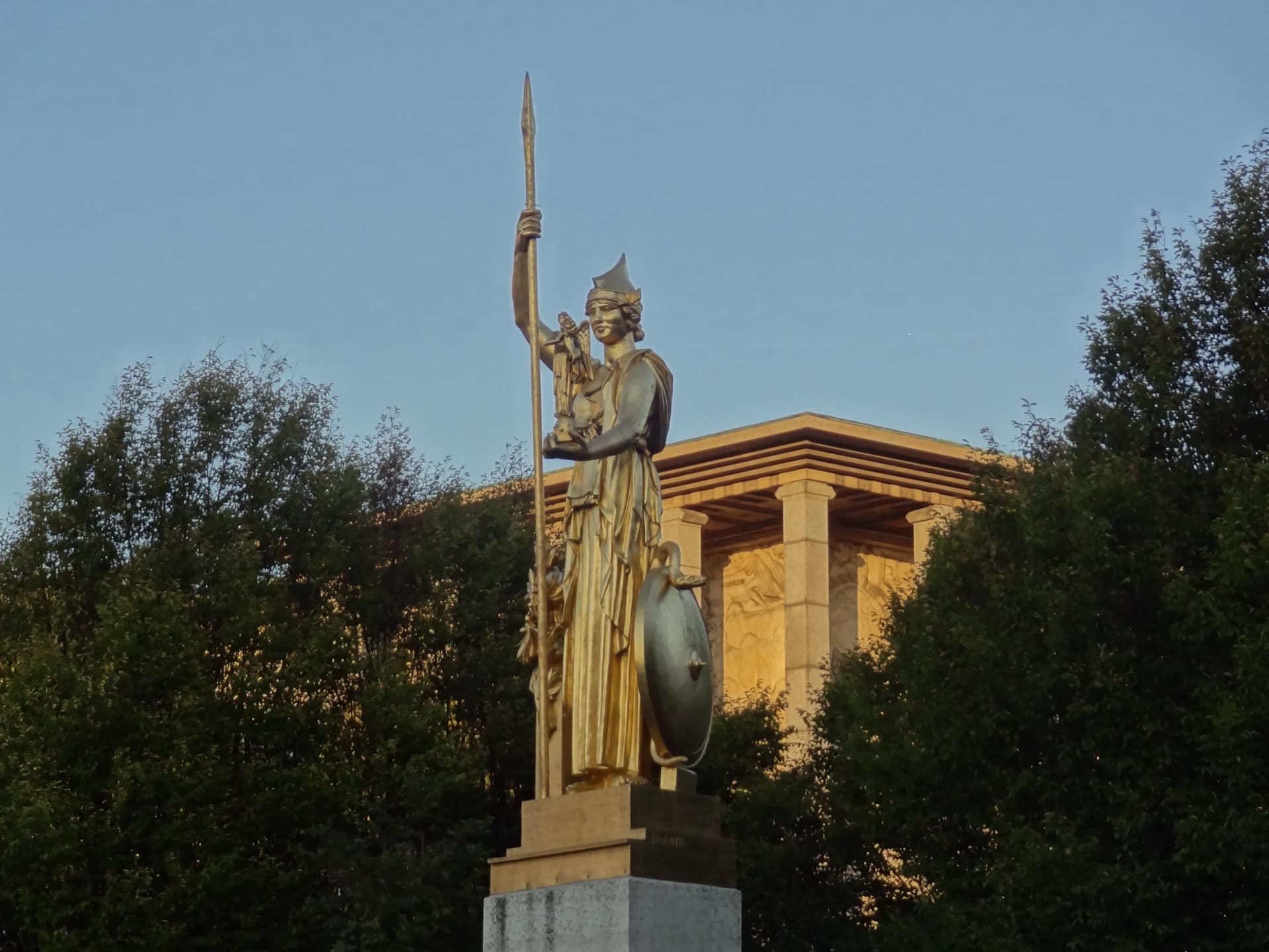 Une statue dorée de femme casquée à la lance et au bouclier devant le Palais de la Porte Dorée, dont on aperçoit la colonnade et le bas-relief sculpté au coucher du soleil
