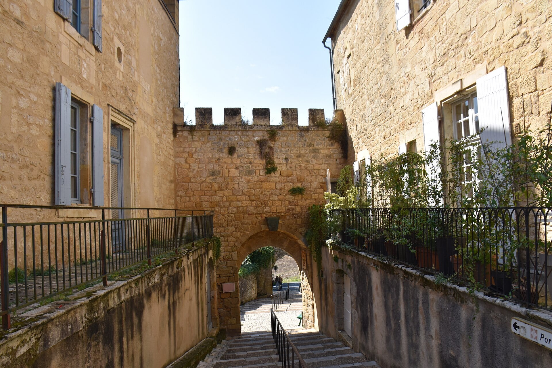 Porte fortifiée en arc de plein cintre surmontée d’un parapet crénelé, encadrée de deux maisons en pierre, avec un escalier menant à l’ouverture.