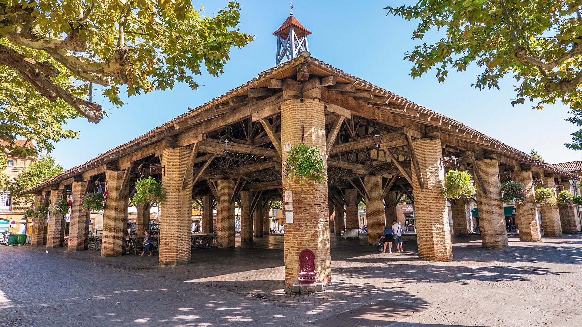 Halle en pans de bois sur piliers de brique, surmontée d’un clocheton, avec jardinières fleuries suspendues et quelques passants sous la charpente.