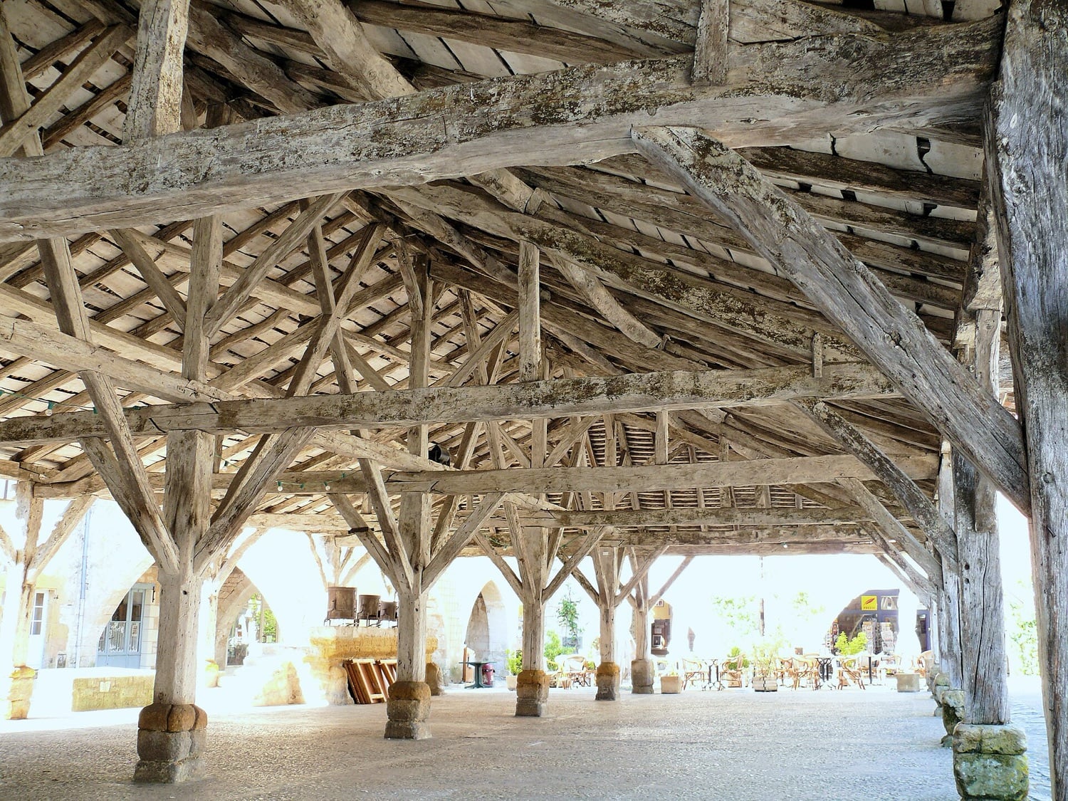 Sous la halle de Monpazier : charpente de bois médiévale à chevrons et entraits, portée par des piliers de pierre à base moulurée, au-dessus d’un sol dallé