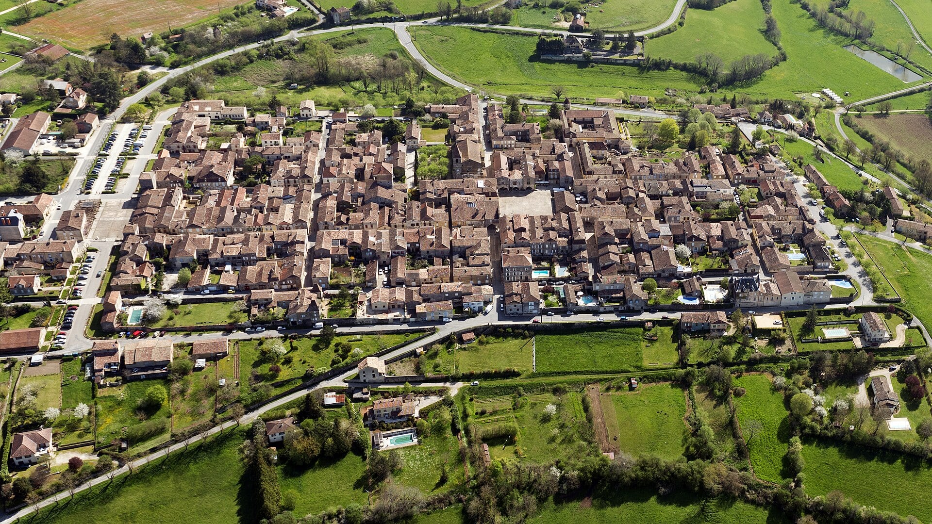 Vue aérienne d’un village organisé selon une trame orthogonale régulière de rues parallèles et perpendiculaires, autour d’une place centrale, entouré de champs et de prairies.