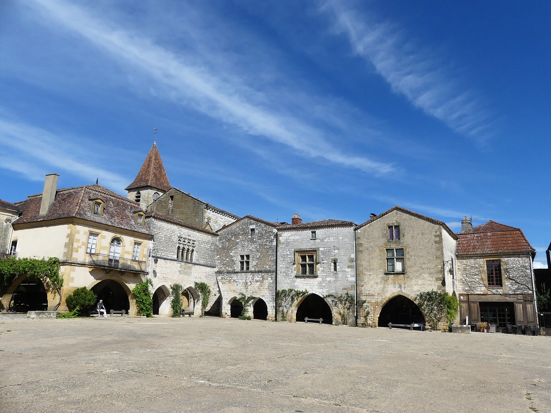 Place bordée de maisons à arcades en pierre (couverts), avec un clocher en arrière-plan et une personne assise sous les arcades.