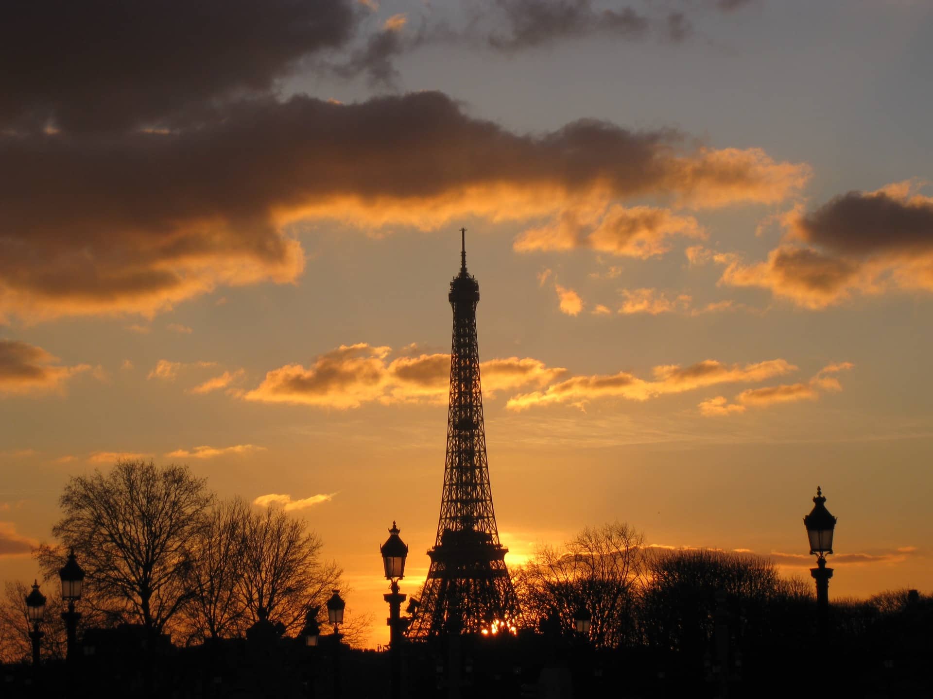 La silhouette de la tour Eiffel se découpant sur un ciel de coucher de soleil orangé, réverbères et arbres nus en ombre au premier plan