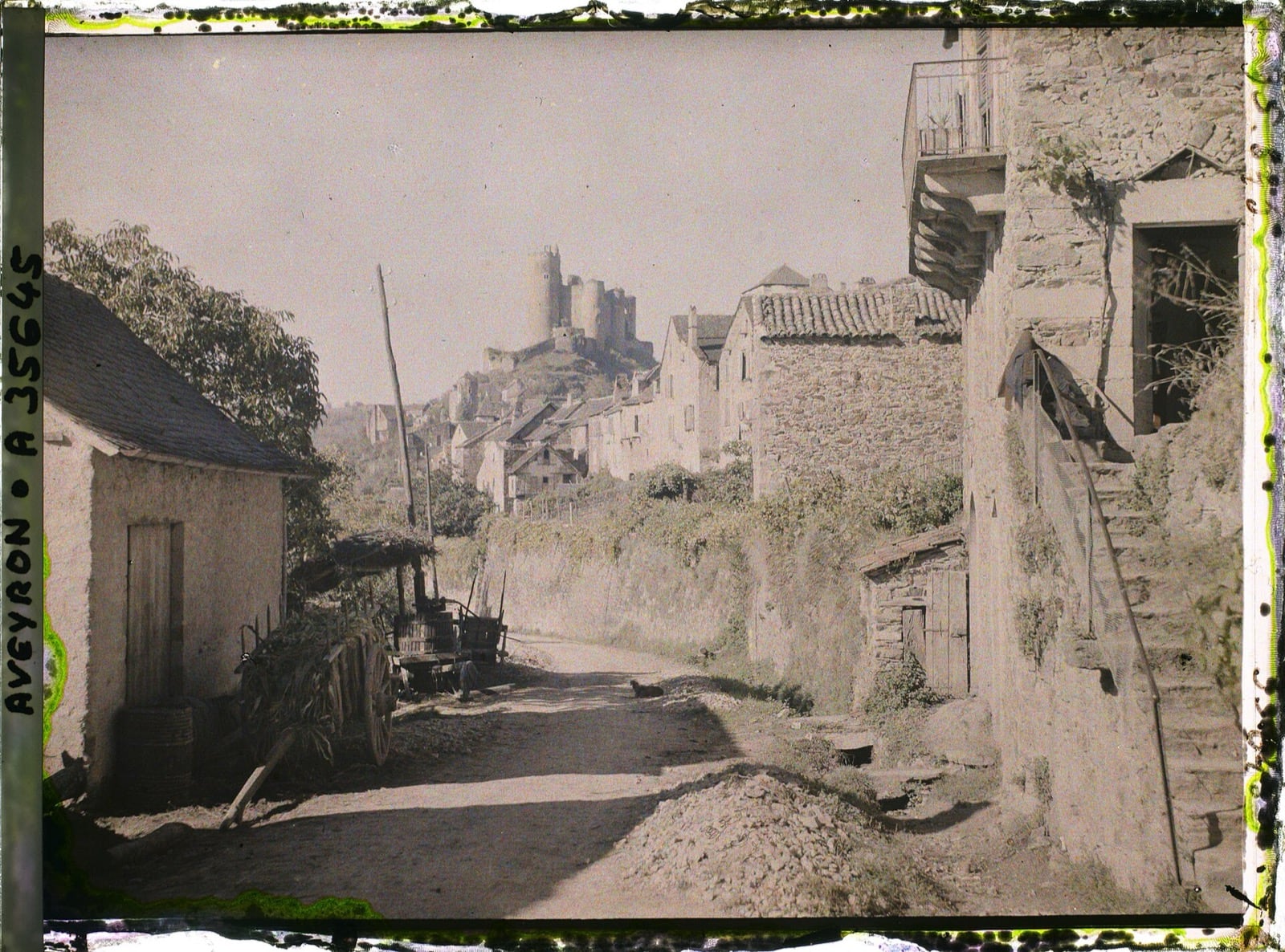 Rue de terre de Najac montant vers la forteresse, un pressoir et une charrette au premier plan, maisons de pierre à escaliers extérieurs