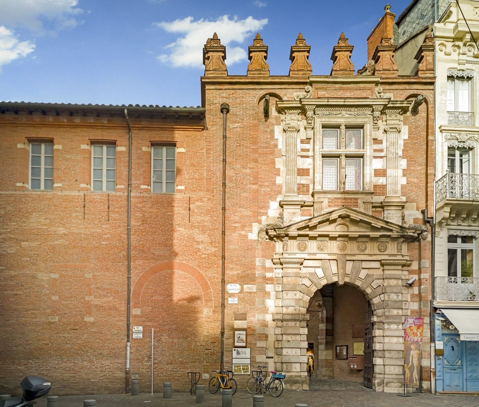 Entrée de l’hôtel d’Assézat sur la place, à Toulouse : haut mur de brique foraine nue percé de fenêtres, portail de pierre à colonnes et fronton, couronnement de brique
