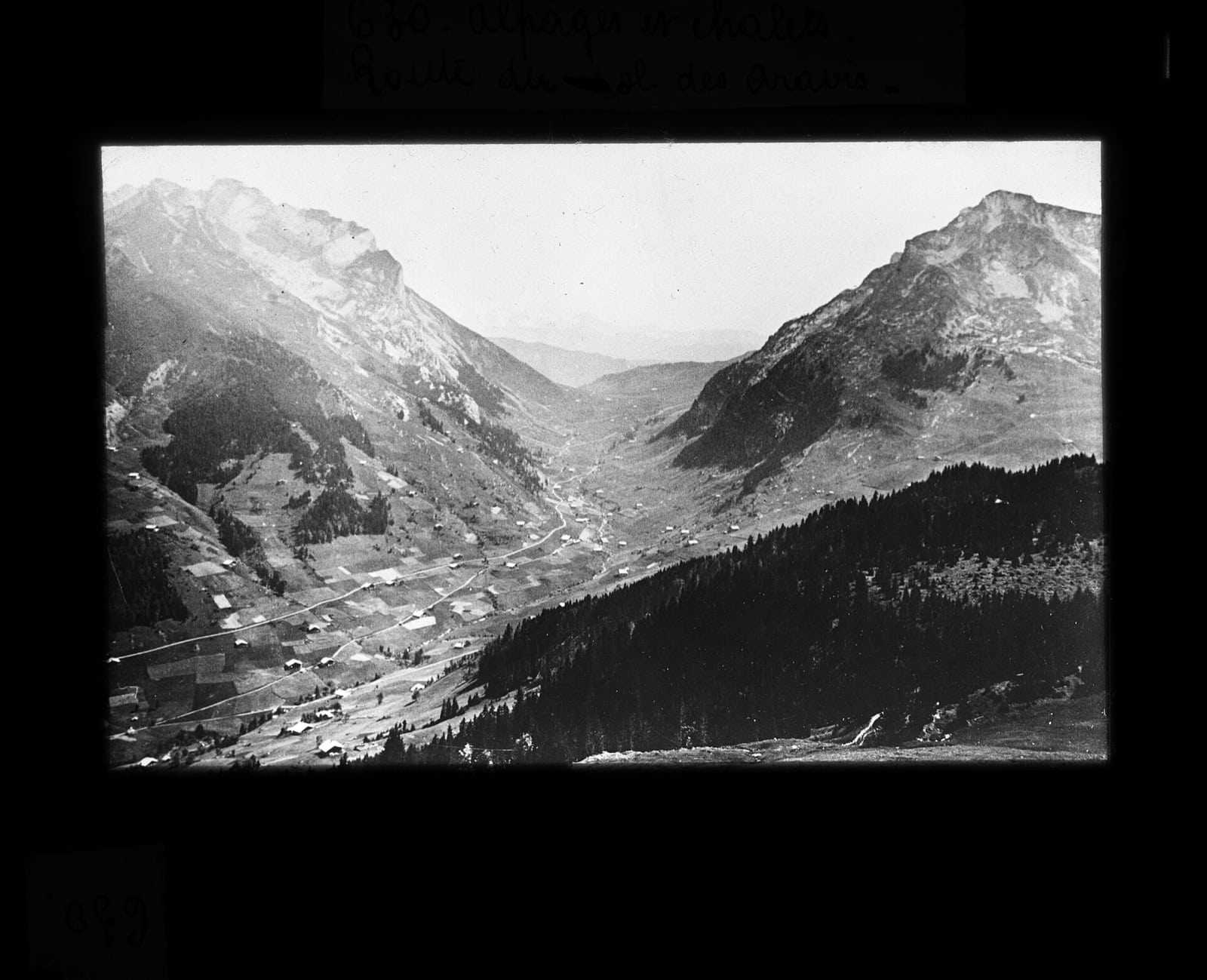 Plaque de projection ancienne montrant une vallée alpine parsemée de chalets et de pâturages, légendée à la main « Alpages et chalets. Route du col des Aravis »