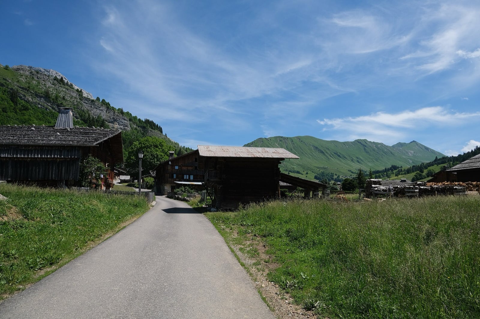 Hameau de chalets et granges de bois sombre le long d’un chemin, dont une grange surélevée à toit débordant, devant les alpages des Aravis au Chinaillon