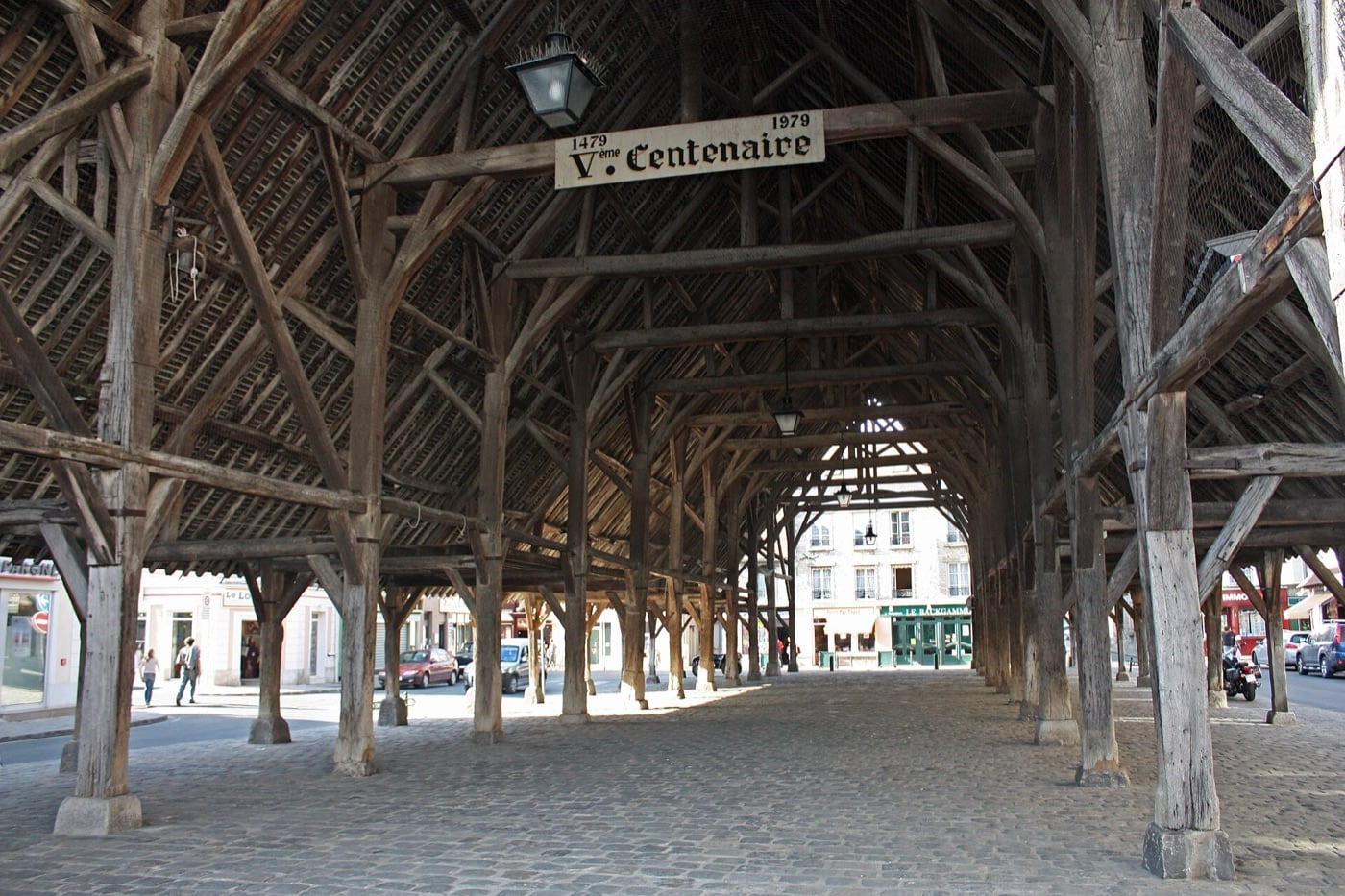 Charpente de la halle de Milly-la-Forêt : forêt de poteaux et de fermes de chêne reposant sur des socles de pierre, sous une vaste couverture.