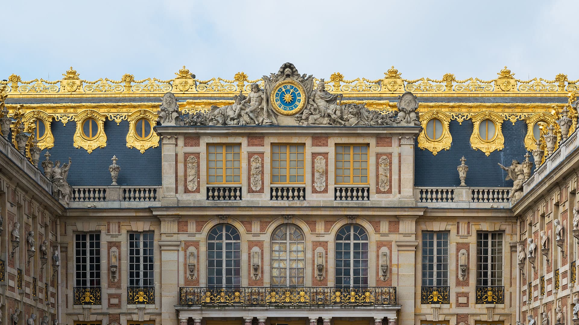 Façade de la cour de Marbre à Versailles : murs de brique et de pierre, toit d’ardoise à œils-de-bœuf, crête de plomb doré, horloge bleu et or entourée de figures allégoriques