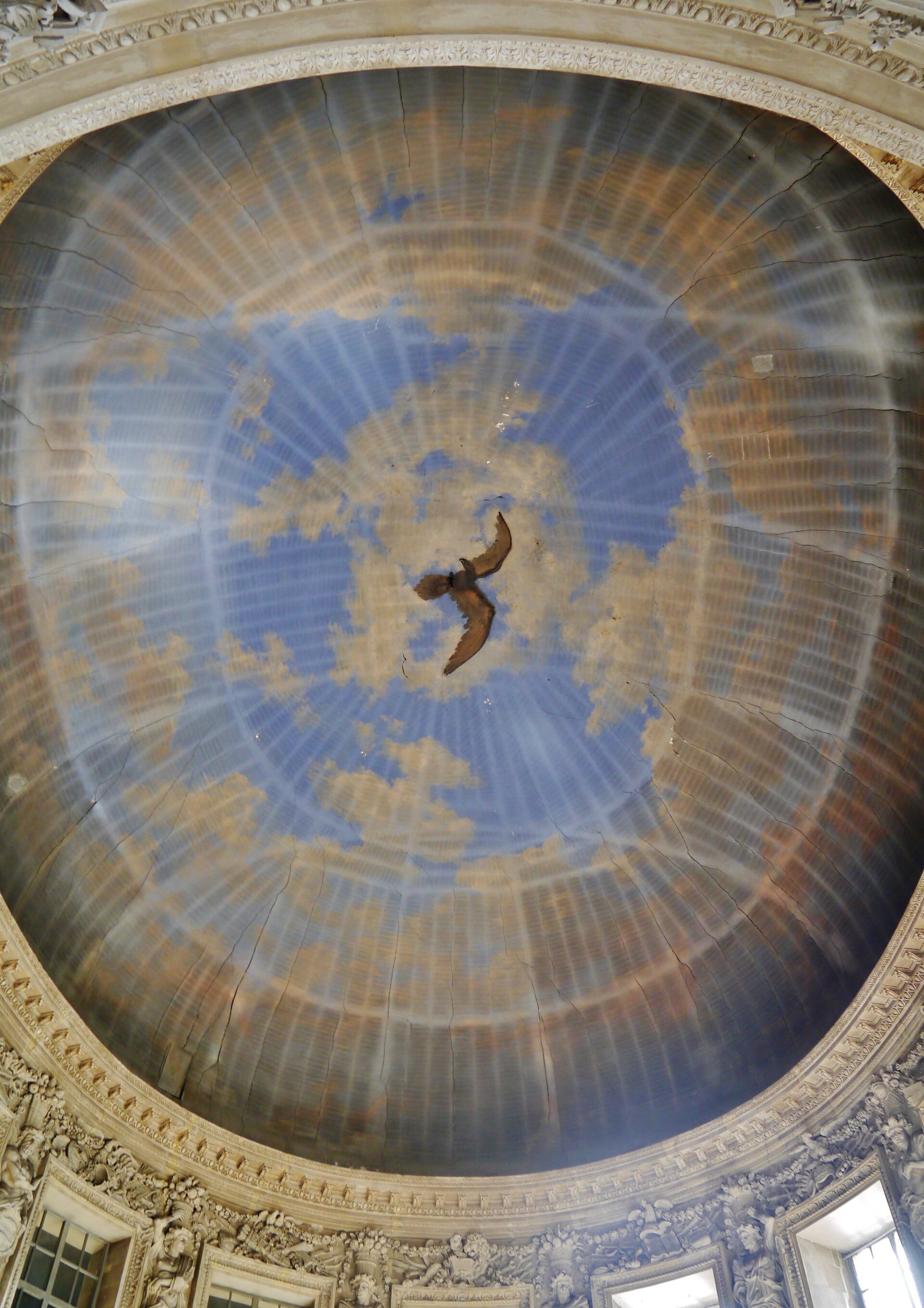 Coupole ovale du Grand Salon de Vaux-le-Vicomte vue de dessous : ciel peint en trompe-l’œil avec un oiseau et un soleil rayonnant, riche corniche sculptée et pendentifs à figures
