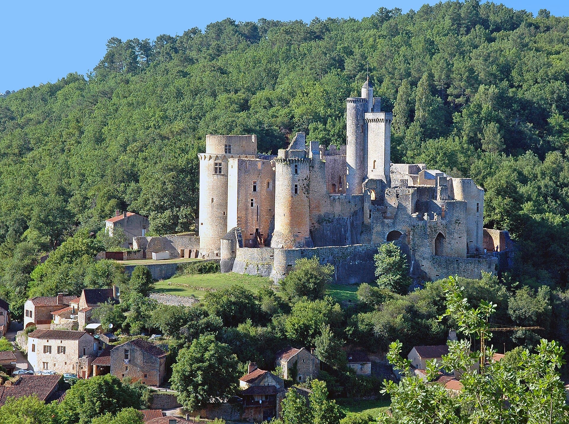 Château de Bonaguil, forteresse ruinée sur un socle rocheux au-dessus d’un village : grosses tours cylindriques, haute tour maîtresse à couronnement crénelé, dominant les toits