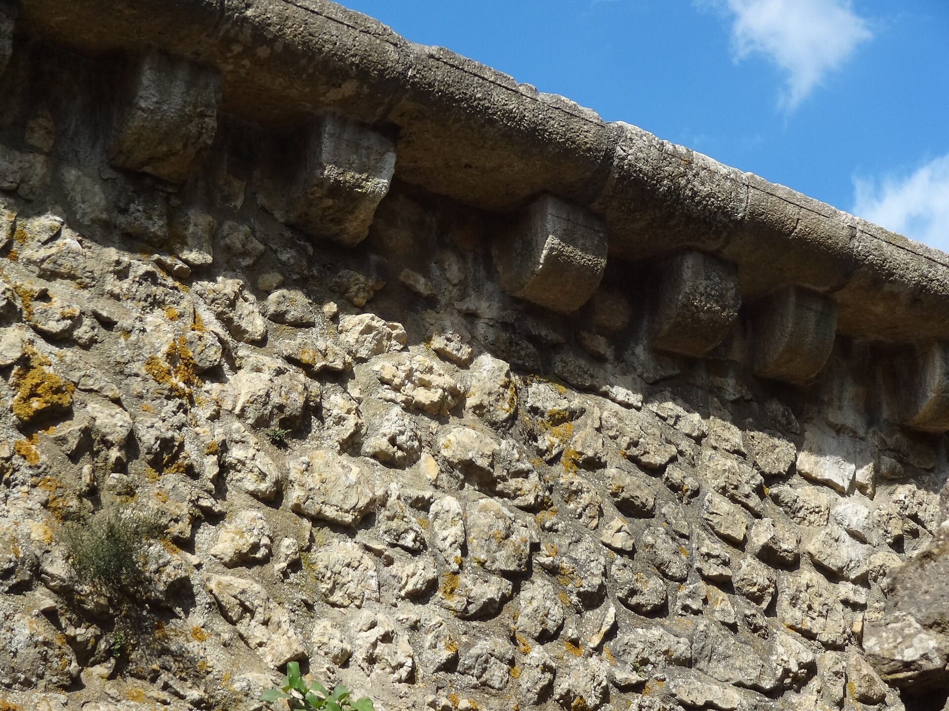 Détail en gros plan du couronnement d’un mur : rangée de consoles de pierre en encorbellement portant un parapet, maçonnerie de moellons en dessous