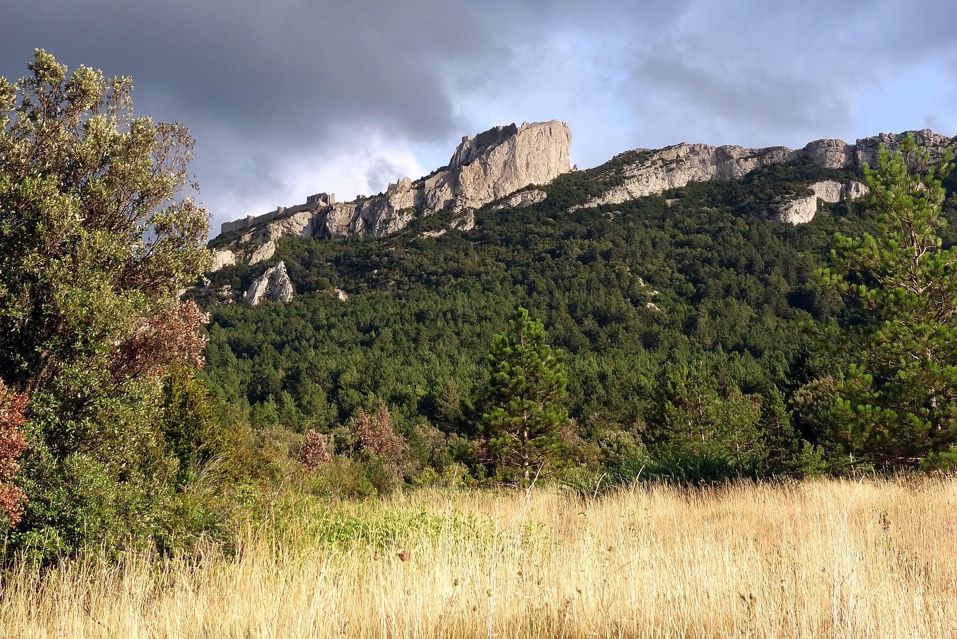 Longue forteresse de pierre épousant la crête d’une montagne calcaire, un donjon dressé sur le point culminant, sous un ciel d’orage