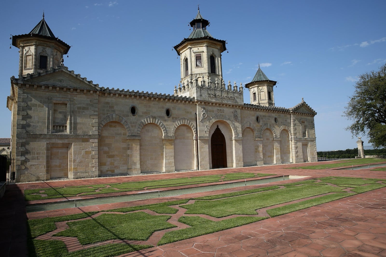 Long bâtiment de pierre claire à arcades, couronné de tours coiffées de toits en pagode et d’une porte en arc brisé, précédé d’un parvis dallé