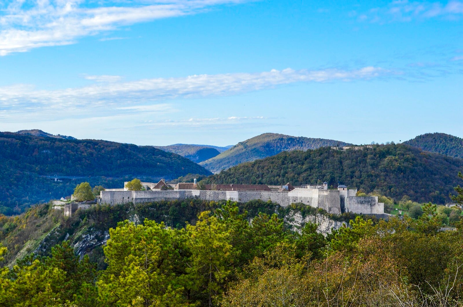 La citadelle de Besançon vue de loin, longue muraille de pierre grise posée au bord d’un plateau rocheux dominant la vallée du Doubs, sommets boisés à l’arrière-plan.