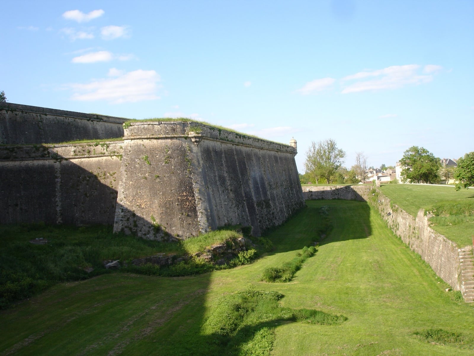 Rempart bastionné de la citadelle de Blaye : haut mur de pierre à flancs inclinés, terre-plein engazonné au sommet, fossé sec en herbe au pied et contrescarpe en face ; une petite échauguette de pierre coiffe l’angle saillant.