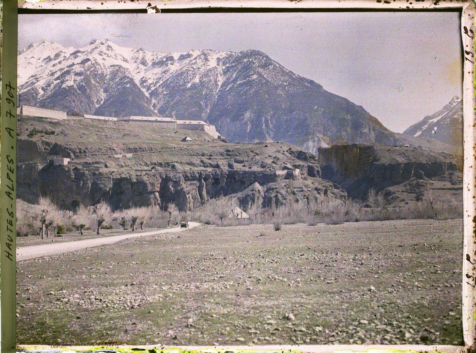 Photographie ancienne en couleurs de Mont-Dauphin : longue muraille blanche de la place forte posée sur un plateau rocheux entaillé par un torrent, hautes montagnes enneigées à l’arrière-plan.