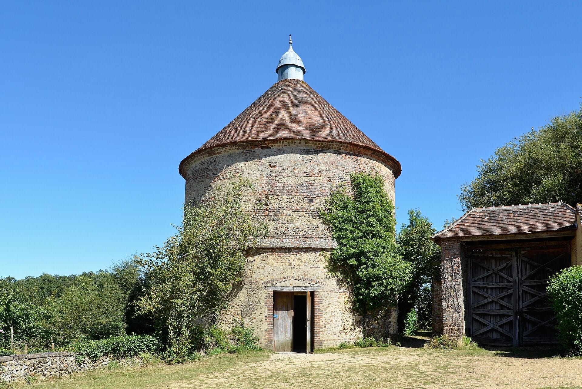 Grand pigeonnier circulaire en pierre et brique coiffé d’un toit conique de tuiles surmonté d’un lanternon de zinc, porte basse à fronton ; à droite une grange à porte charretière de bois, cour d’herbe et muret de pierre sèche sous un ciel bleu