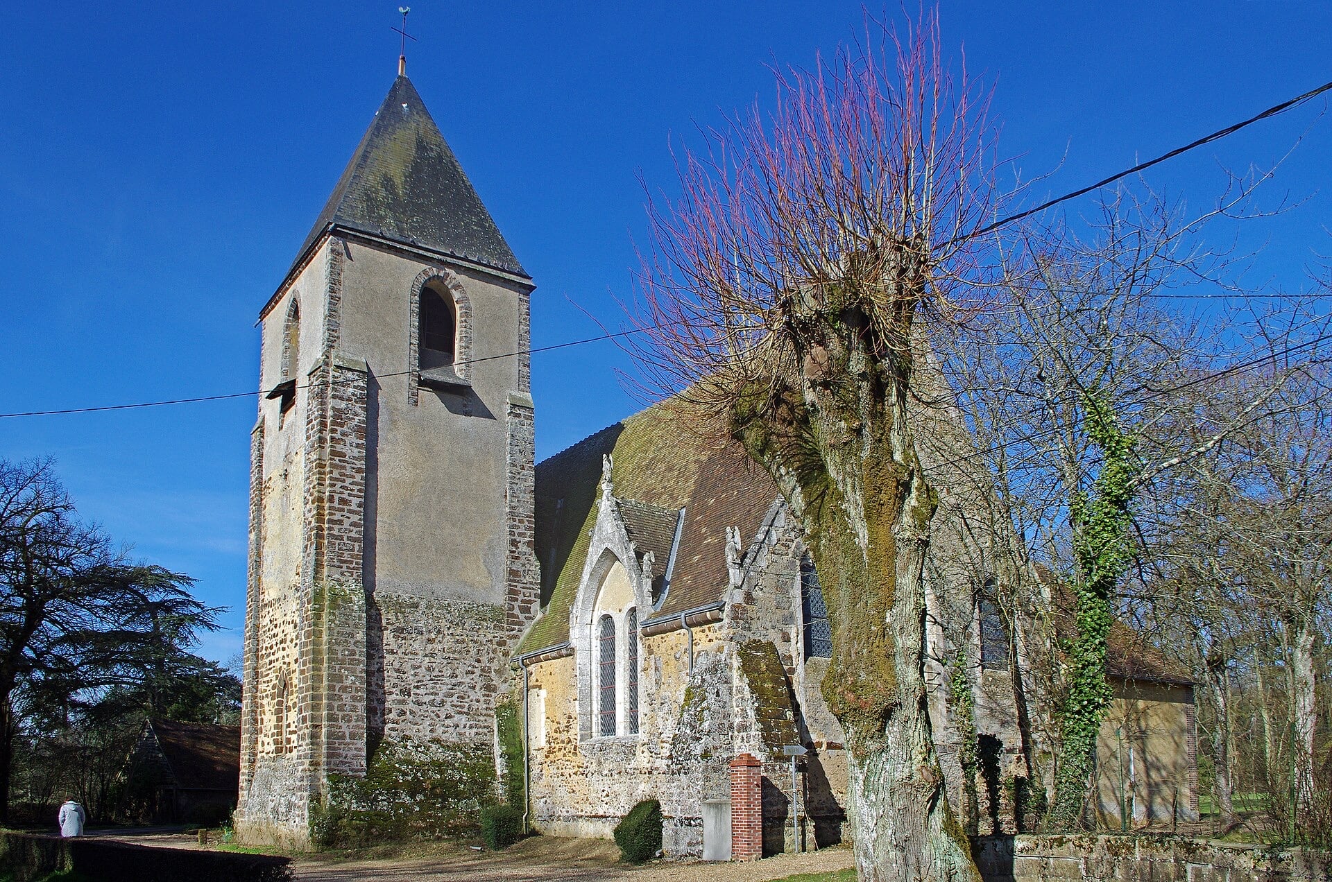 Église rurale au clocher-tour carré coiffé d’une flèche d’ardoise, nef basse à toit de tuiles se terminant par un mur droit sans abside, contreforts et lucarne à gable, grand arbre têtard au premier plan sous un ciel bleu