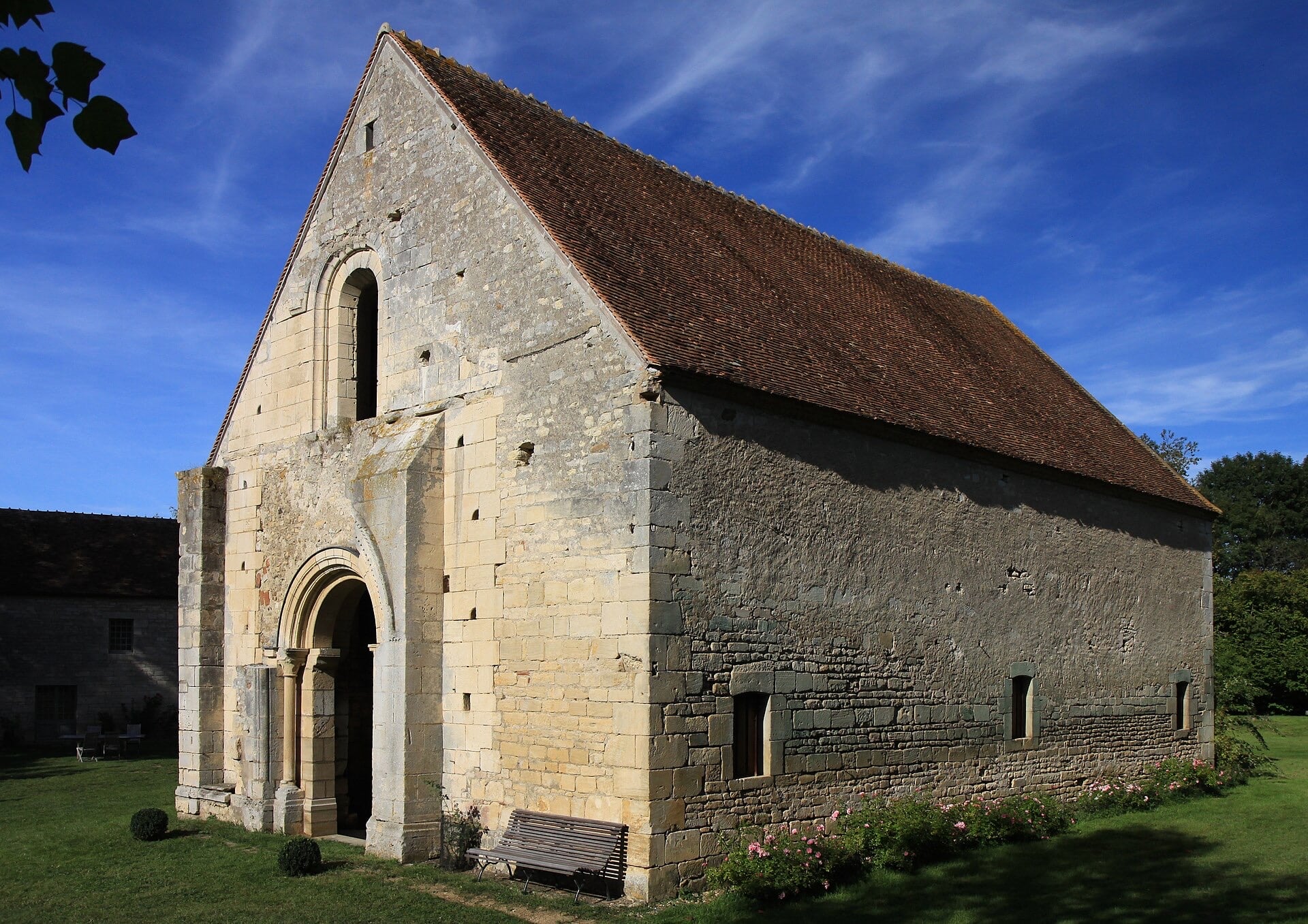 Chapelle rurale en pierre à nef unique et haut toit de tuiles à forte pente : pignon percé d’une baie en plein cintre au-dessus d’un portail roman à voussures, longue façade latérale à contreforts et petites fenêtres étroites, pelouse et rosiers au pied
