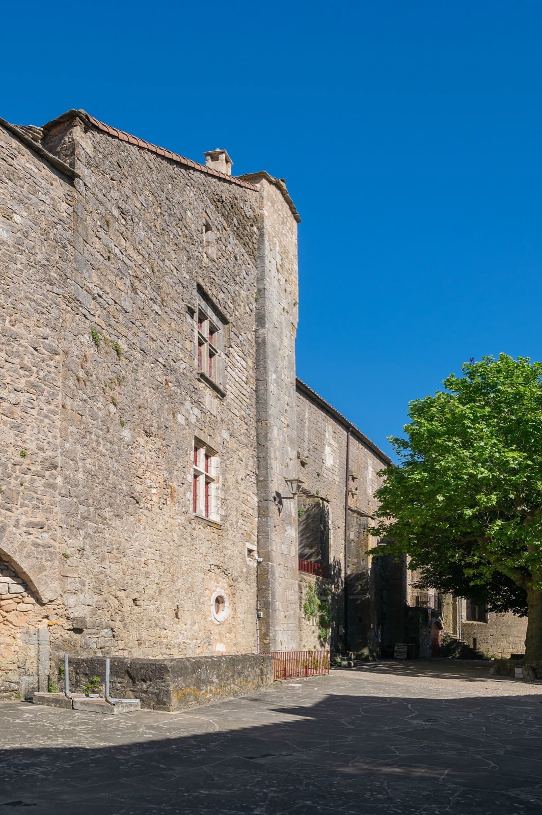 Haut bâtiment médiéval en pierre calcaire sur une place pavée : façade percée de fenêtres à croisée aux volets rouges et d’un petit œil-de-bœuf, chaînes d’angle, toit de tuiles ; à droite un platane et l’enfilade des maisons du village