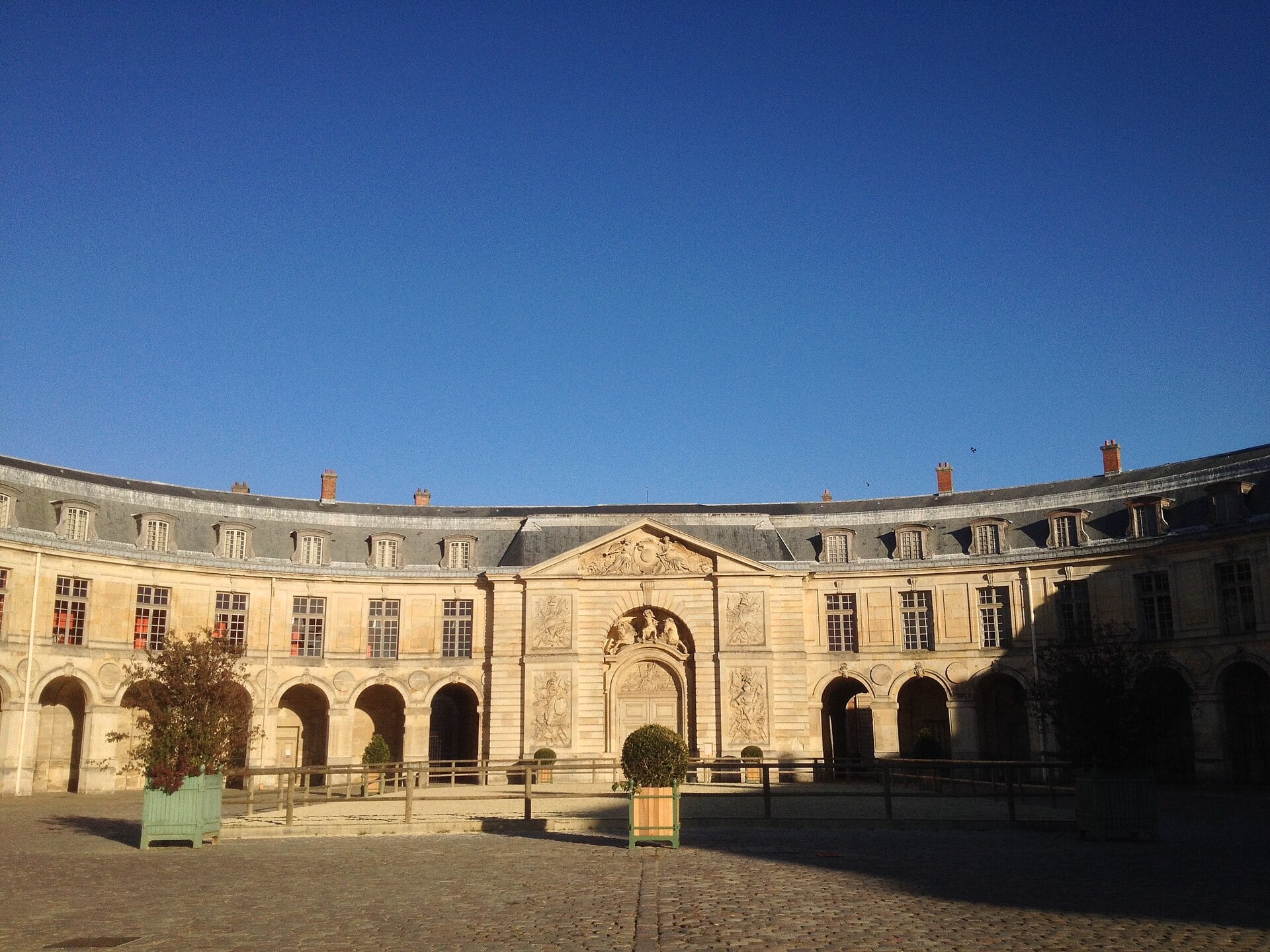 La cour en hémicycle de la Grande Écurie de Versailles : longue façade courbe de pierre de taille rythmée d’arcades, avant-corps central à fronton sculpté percé d’un grand portail cintré, lucarnes en toiture.