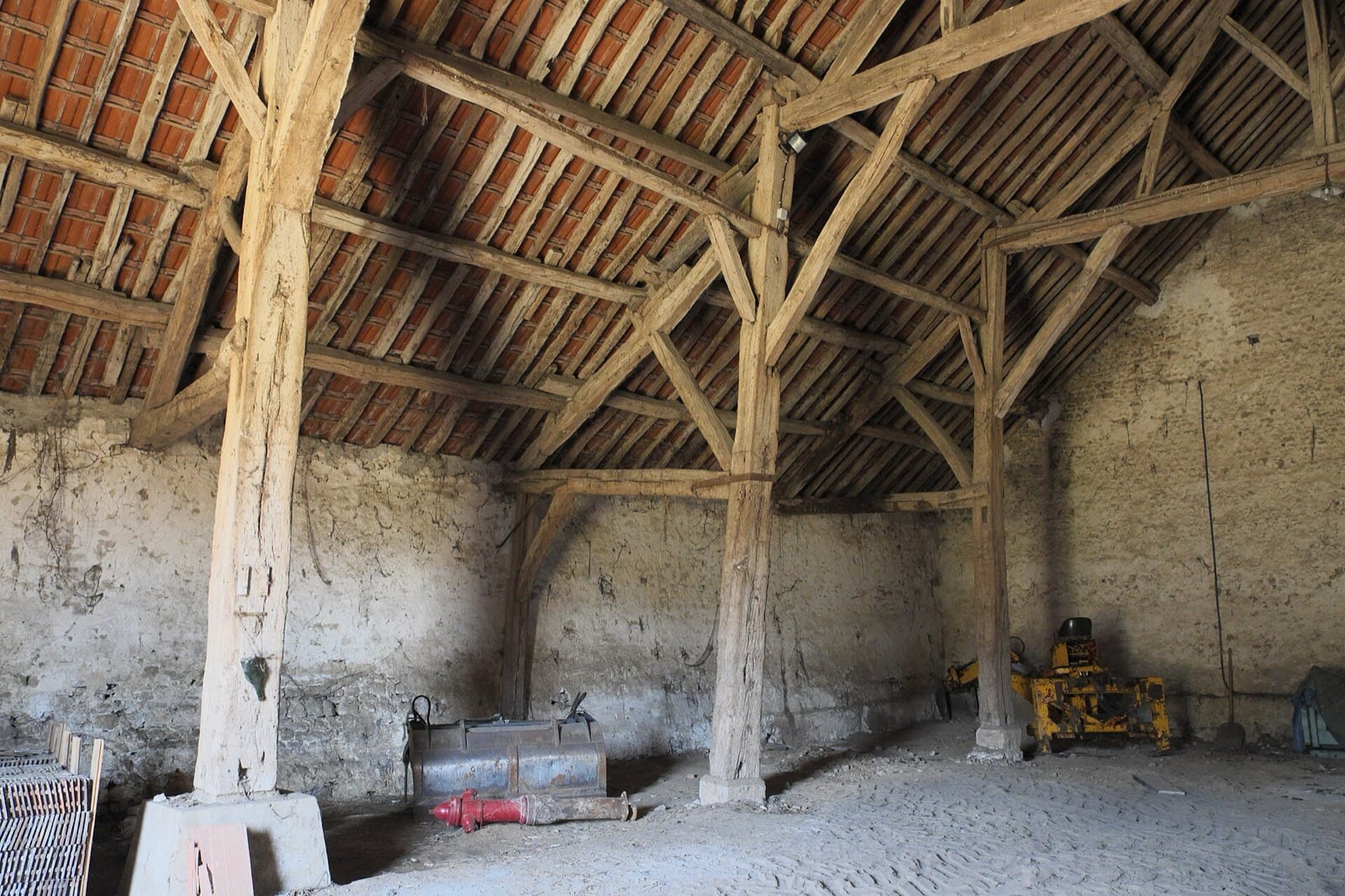 Intérieur d’une grange : hauts piliers de bois dressés sur des dés de pierre divisant le volume en vaisseaux, portant une vaste charpente de chêne sous une couverture de tuiles.