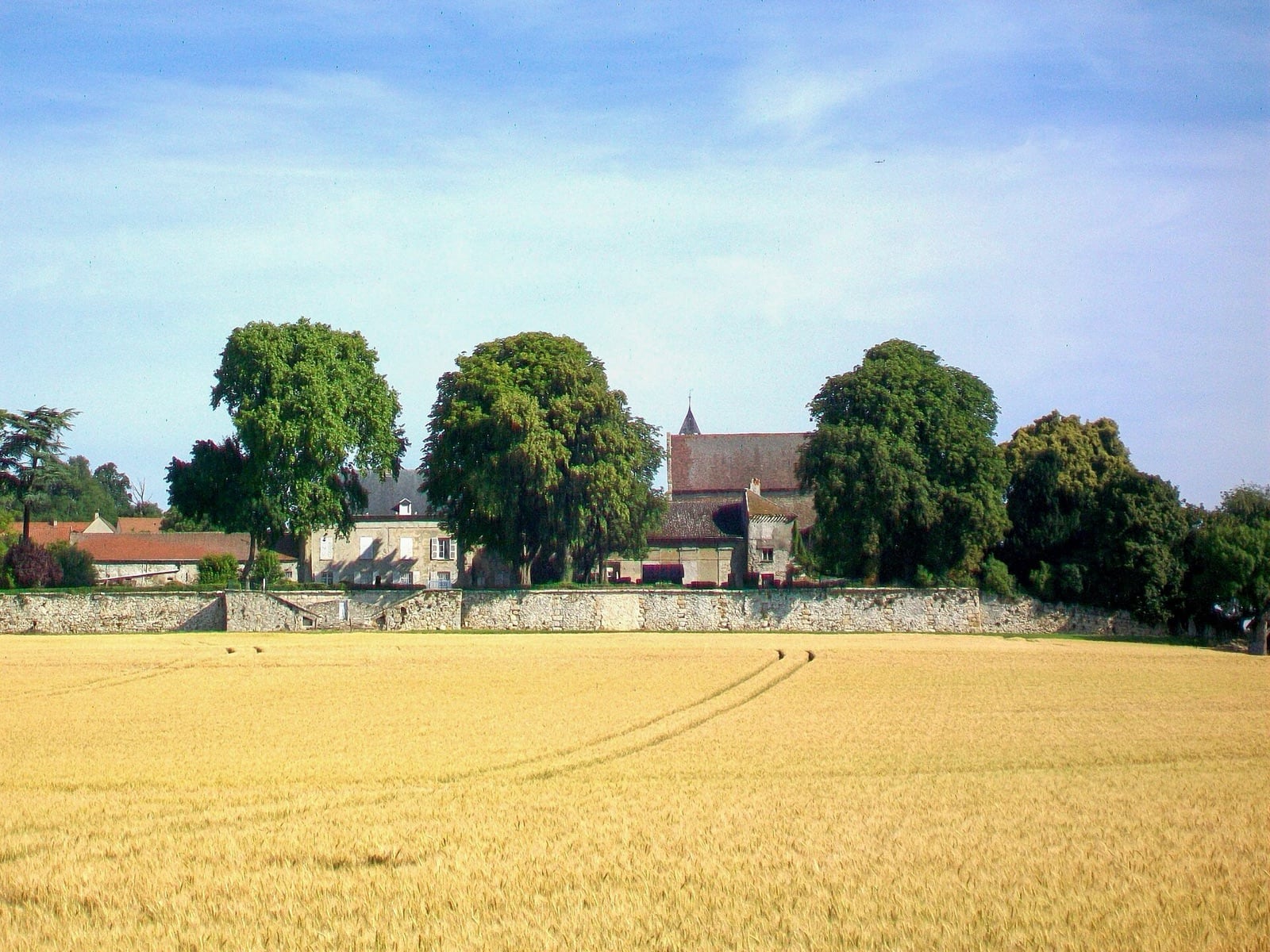 Grande ferme et son manoir enclos derrière un long mur de pierre, dominés par un clocher et de grands arbres, au bord d’un champ de blé mûr traversé de traces de roues.