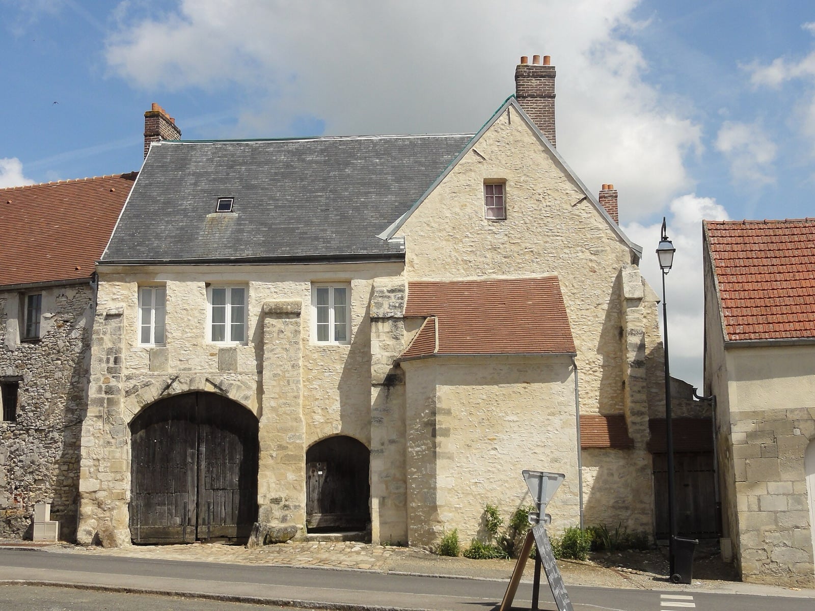 Porche de ferme en pierre de taille : grande arche charretière fermée de vantaux de bois et arche piétonne plus étroite, surmontées d’un logis à comble et cheminées, sous un toit d’ardoise.
