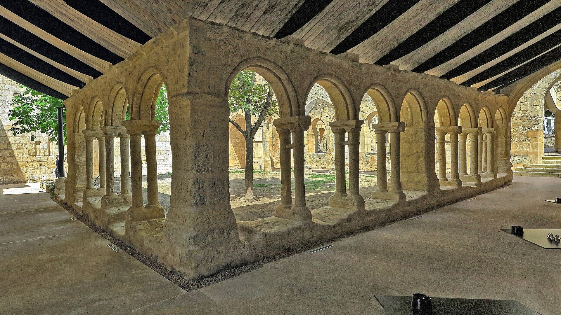 Galerie de cloître en pierre calcaire blonde à colonnettes jumelées monolithes et arcs brisés, ouverte sur un jardin planté d’un arbre, sous une couverture moderne à poutres
