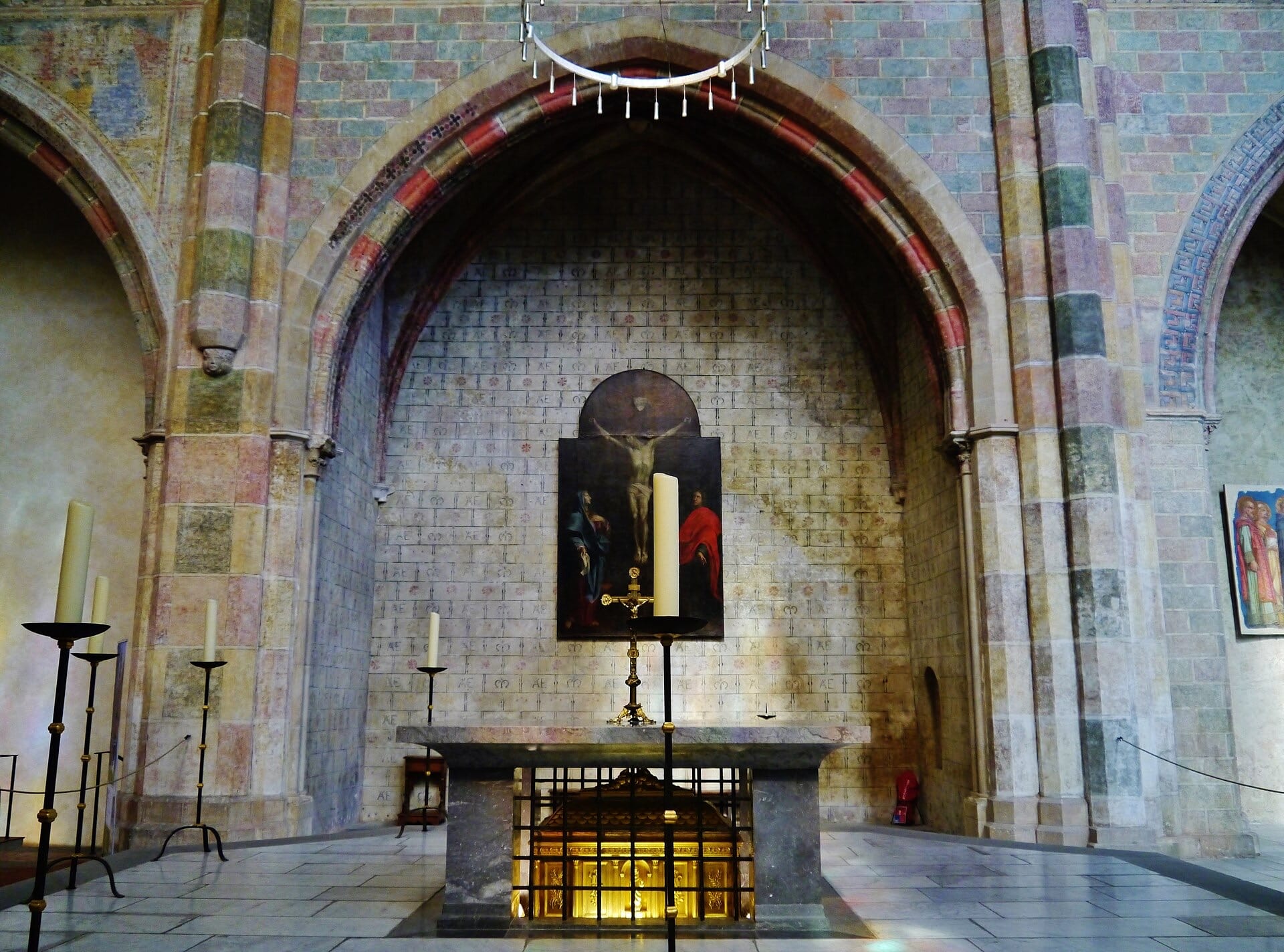 Travée d’église gothique aux murs de brique peints de motifs, grande arcade brisée abritant un autel de pierre, tableau d’une Crucifixion et châsse dorée sous l’autel