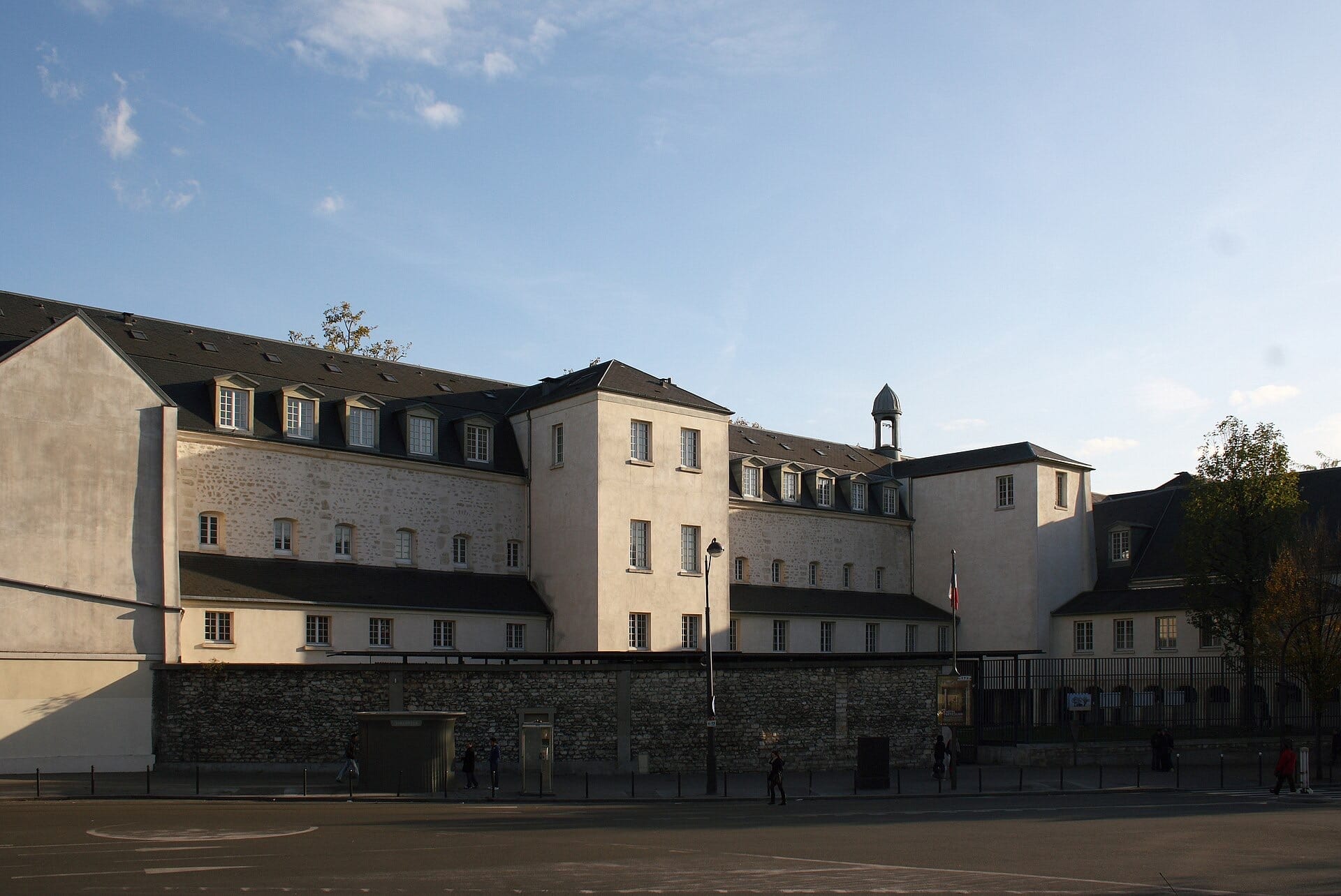 Long bâtiment classique de trois niveaux à façades claires et toit d’ardoise à lucarnes, cantonné de pavillons et surmonté d’un clocheton, précédé d’un mur de clôture en moellon bordant une rue