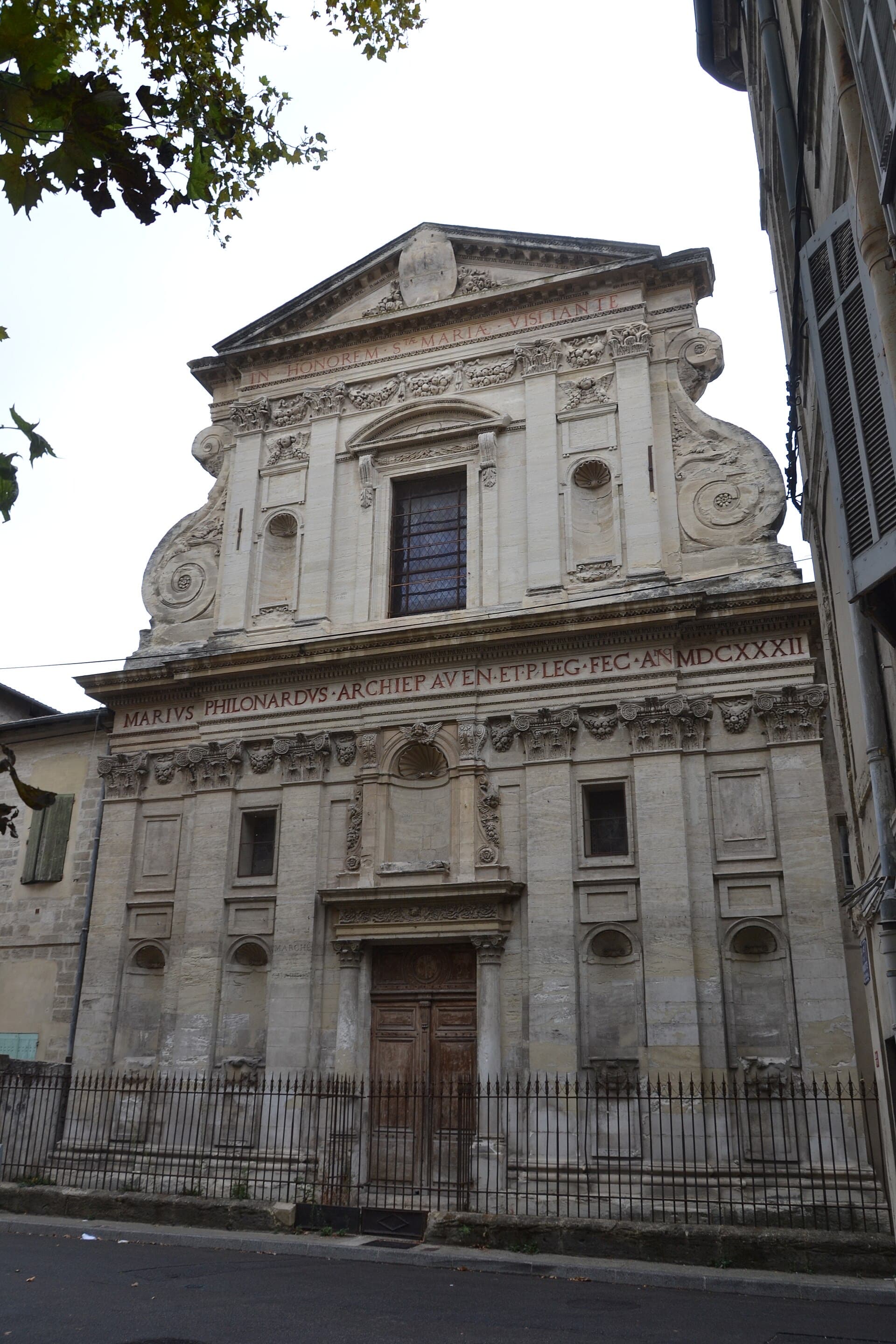 Façade d’église baroque en pierre claire à deux registres, ornée de volutes, pilastres, niches à coquille et frontons, portant deux longues inscriptions latines peintes en rouge, derrière une grille