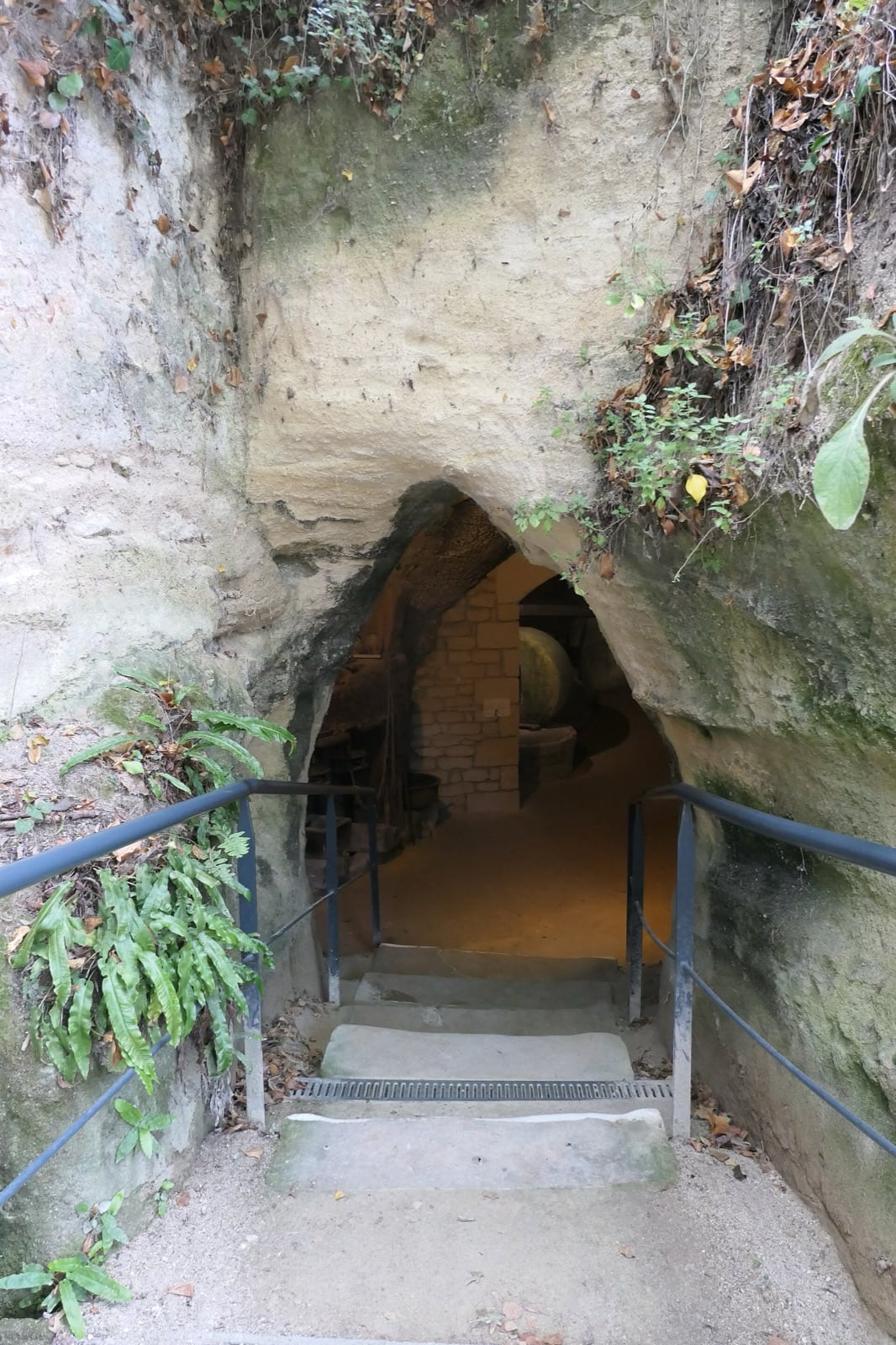 Escalier de pierre descendant sous une arche taillée dans la roche vers l’entrée sombre d’une cave creusée ; à l’intérieur, une chambre voûtée en briques et de vieux ustensiles dans la pénombre.