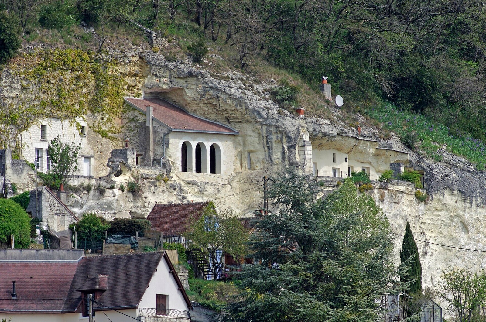 Falaise de tuffeau blanc entaillée de logis : maisons à toits de tuiles adossées au rocher, une façade percée d’une arcade à trois baies cintrées et, plus à droite, des entrées de caves à devantures blanchies ouvertes dans la pierre.