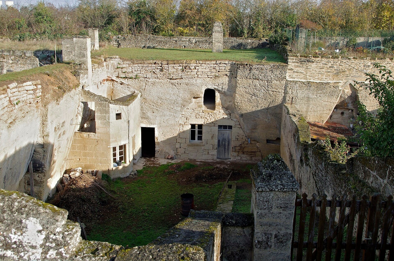 Vue plongeante sur une cour rectangulaire creusée à ciel ouvert dans le falun blanc : au fond de la fosse, un logis à pignon, une porte de bois, des fenêtres et des ouvertures de pièces taillées dans les parois ; au-dessus, un jardin sur le plateau.