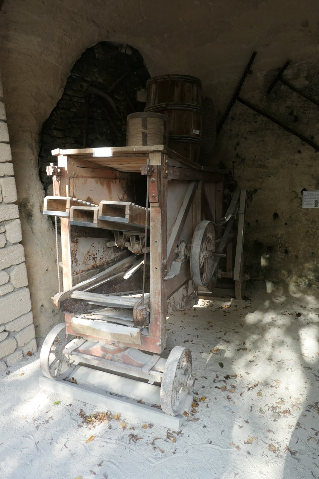 Ancienne batteuse de bois montée sur roues à jantes de fer, avec sa poulie et sa courroie d’entraînement, remisée dans une grange dont les parois et la voûte sont taillées dans le tuffeau ; à gauche, une ouverture cintrée sur un mur de moellons.