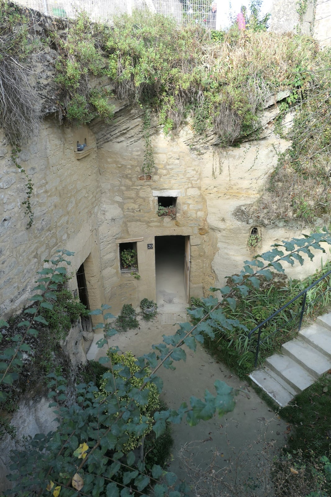 Cour en contrebas creusée dans le rocher : dans la paroi de falun, une porte numérotée « 20 » et des fenêtres encadrées de pierre de taille garnies de géraniums, des niches taillées dans le mur et, à droite, un escalier de pierre descendant dans la fosse.