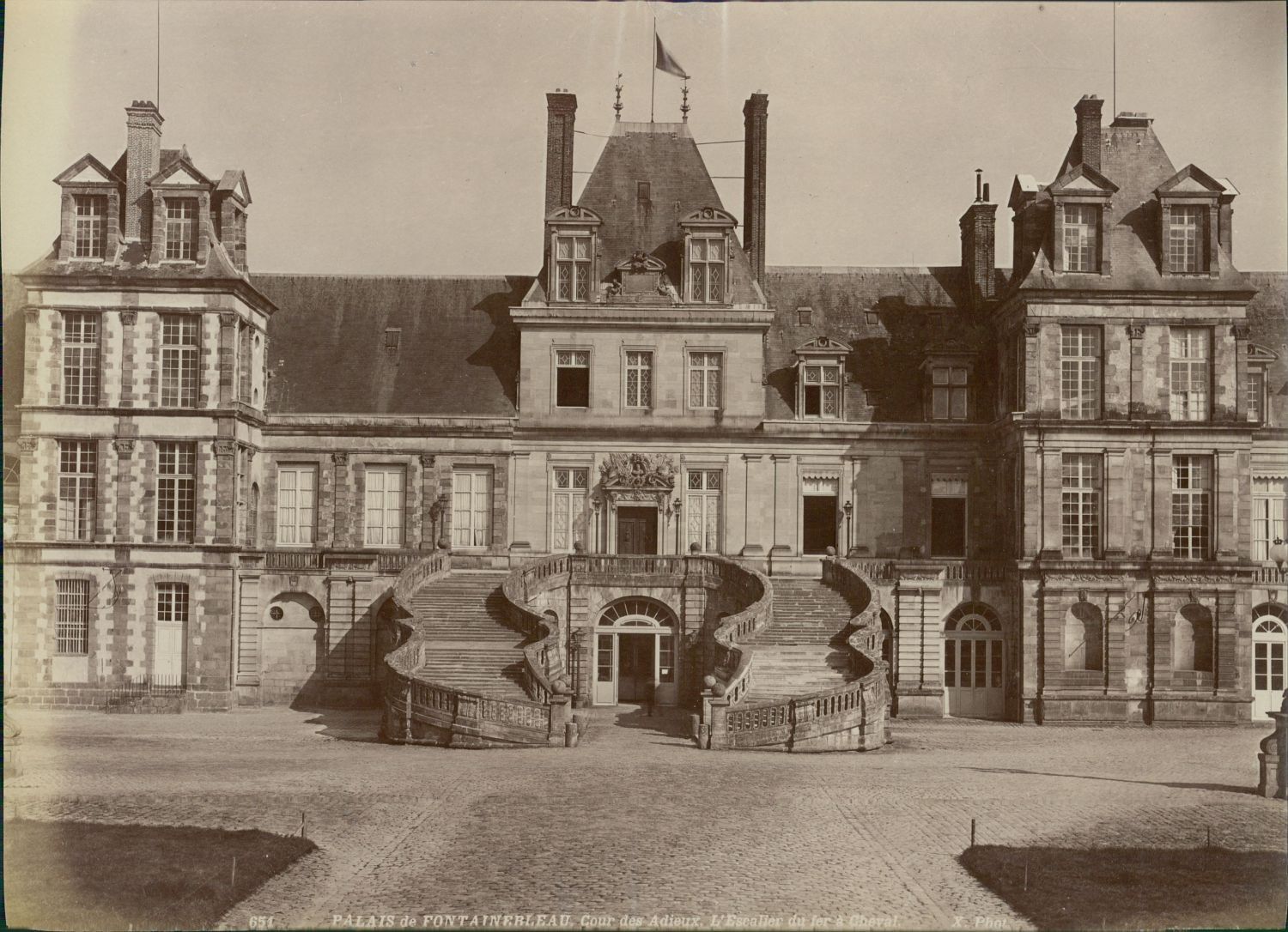 Photographie ancienne de l’escalier en fer à cheval de la cour des Adieux à Fontainebleau : deux volées courbes de pierre se rejoignant sur un palier, en avant-corps de la façade du palais.
