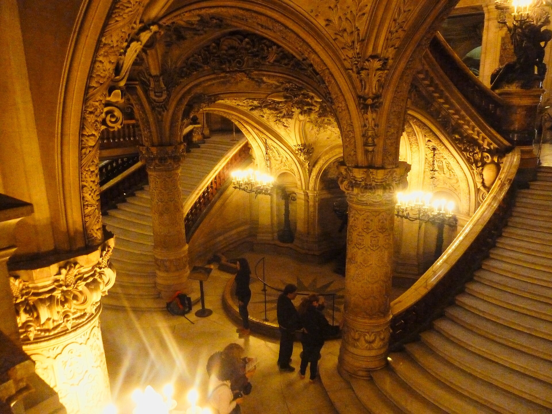 Le grand escalier du palais Garnier vu de côté : volées de marbre montant sous des voûtes sculptées portées par des colonnes de marbre polychrome, éclairées de candélabres.