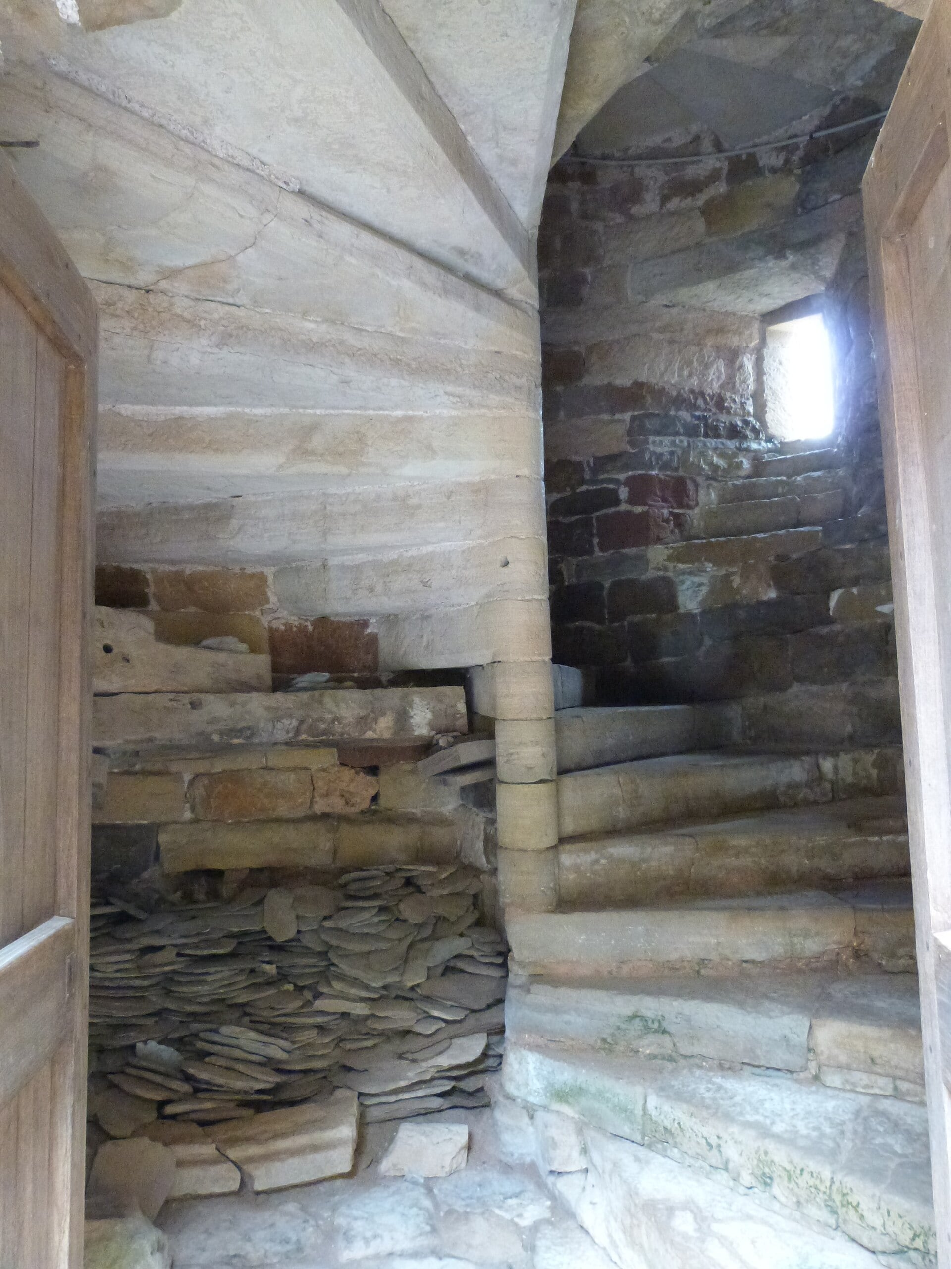 Escalier à vis de pierre d’un château médiéval : marches rayonnantes usées tournant autour d’un noyau central, dans une cage étroite éclairée d’une étroite baie.