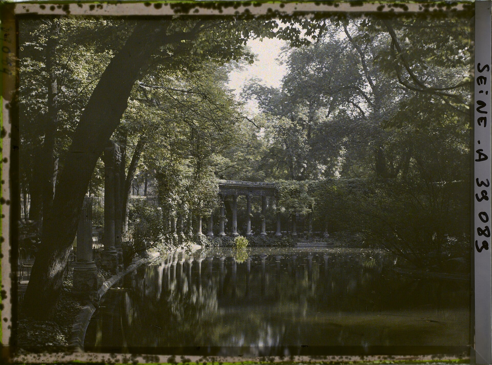 Autochrome ancien de la Naumachie du parc Monceau à Paris : colonnade antique au bord d’un bassin, doublée de son reflet dans l’eau calme, sous un couvert d’arbres.
