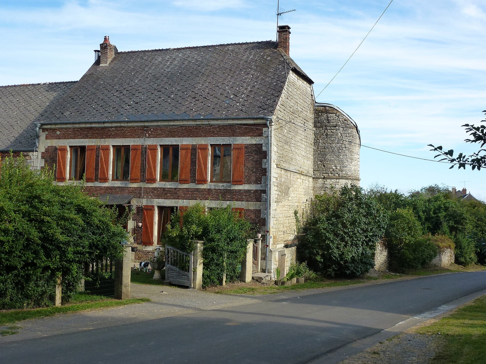 Ferme en brique et pierre à toit d’ardoise, dont l’angle est occupé par une grosse tour ronde en pierre calcaire, en bordure d’une rue de village