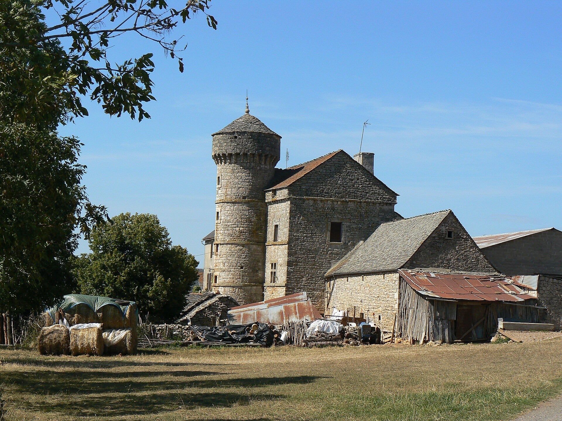 Ensemble rural en pierre calcaire dominé par une haute tour ronde couronnée d’un rang de mâchicoulis et coiffée d’un toit conique en lauzes, accolée à un logis à haut pignon, avec granges et hangars agricoles autour