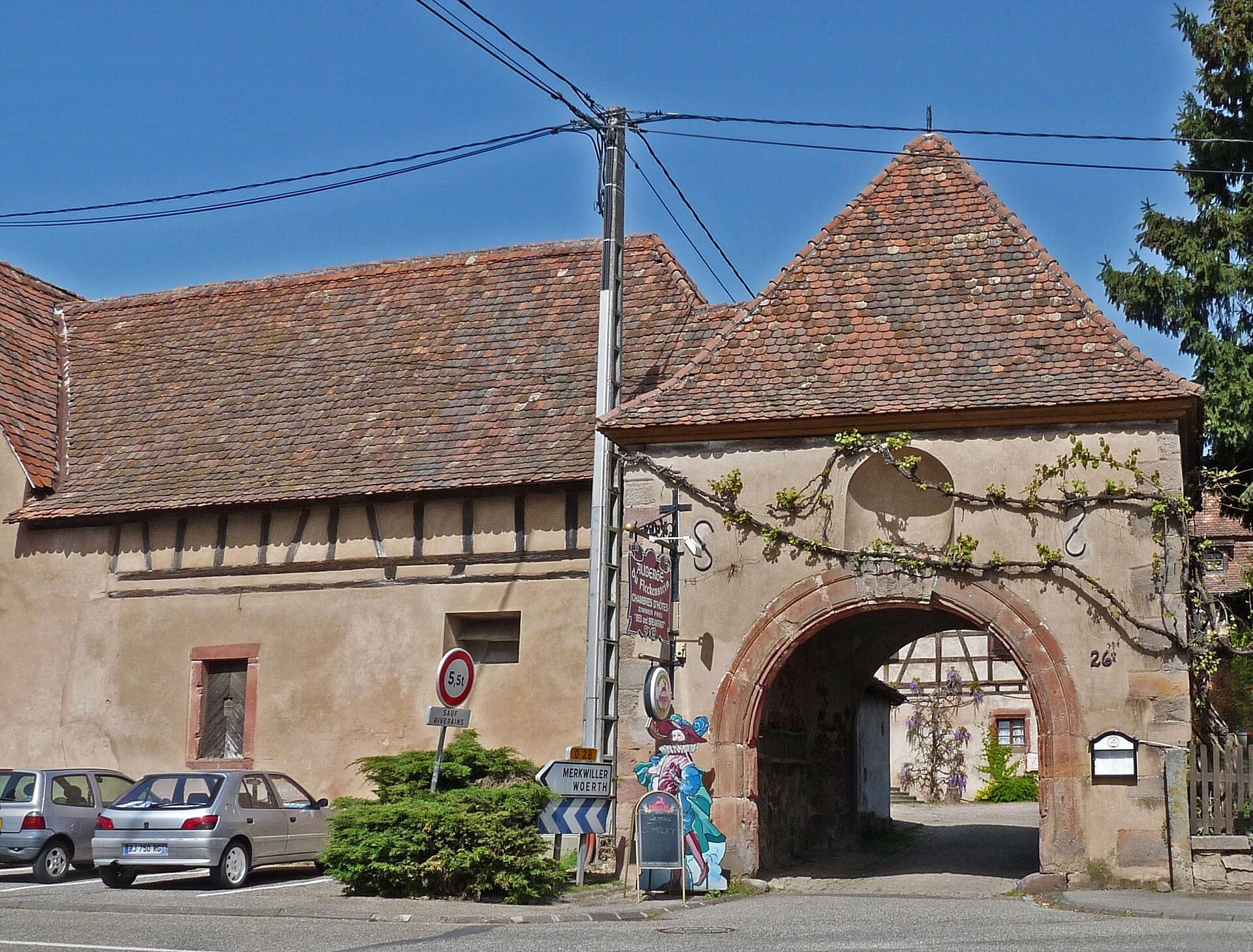 Ancienne ferme alsacienne : haut porche voûté en grès rouge à toit de tuiles ouvrant sur une cour intérieure où se voit une aile à colombage, accolé à une grange de pierre et de pan de bois